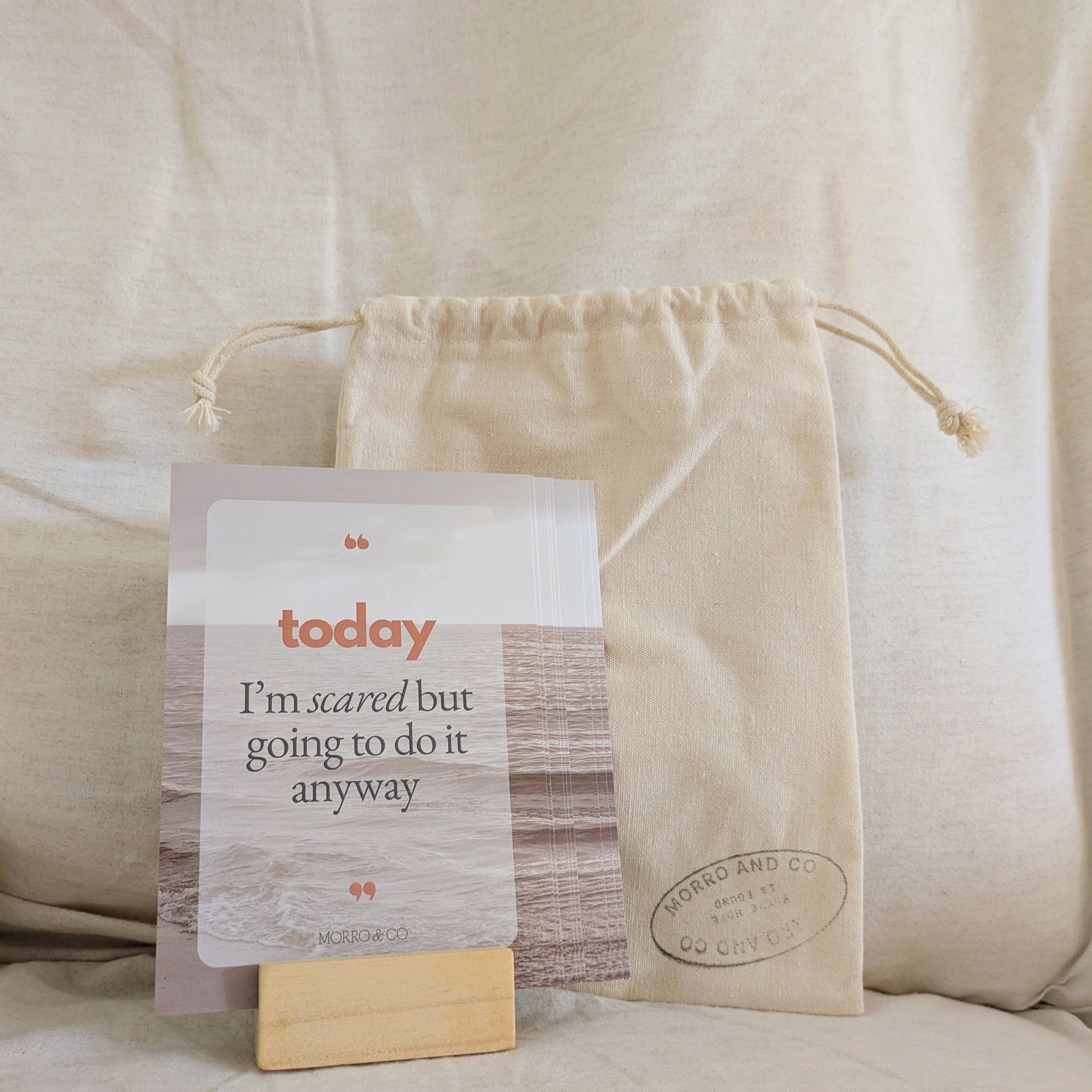 A set of Morro & Co brand affirmation cards displayed on a wooden stand with a positive message on the front, placed next to a beige pouch.