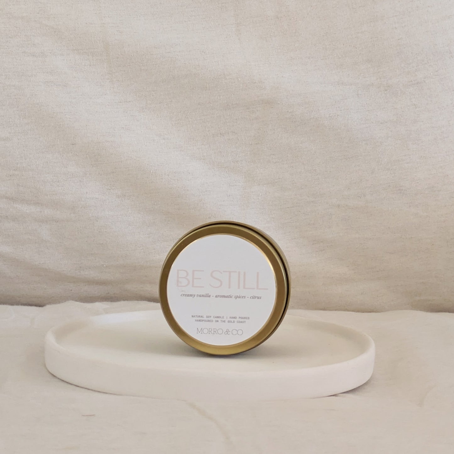 Gold candle travel tin with 'Be Still' text on a beige surface