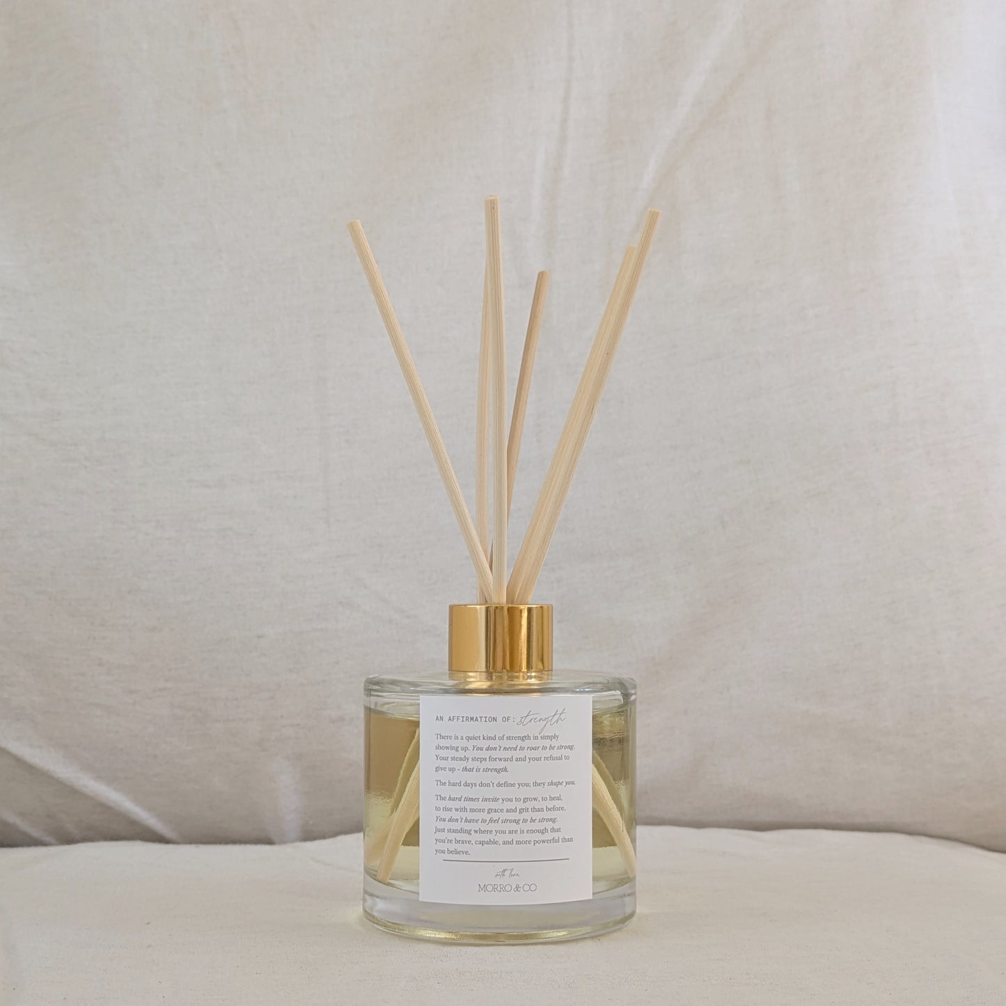 Eco reed diffuser with gold cap, affirmation label and clear bottle on a neutral background