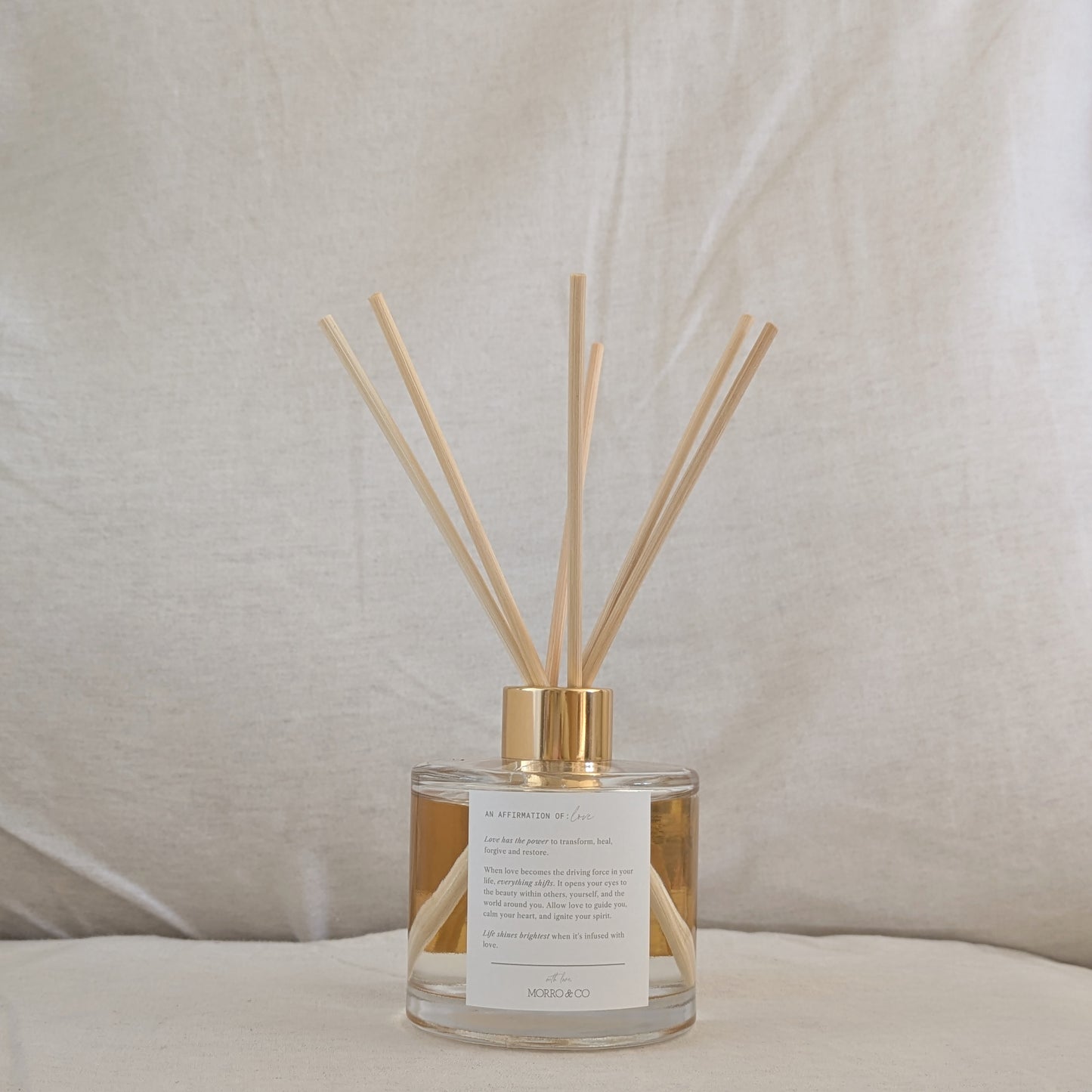 Reed diffuser with gold cap, affirmation label and clear container on a neutral background