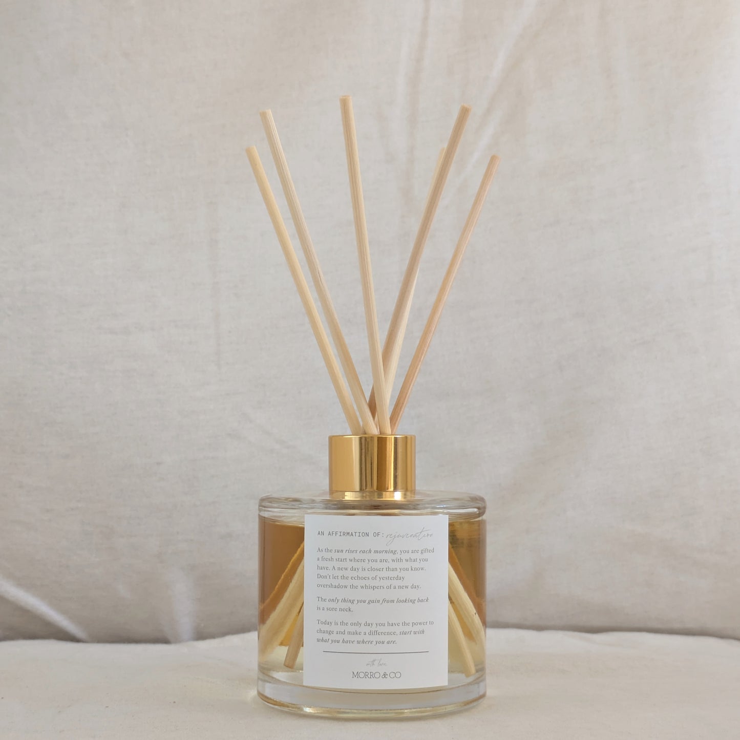 Natural eco reed diffuser with gold cap and clear container, affirmation label on a neutral background