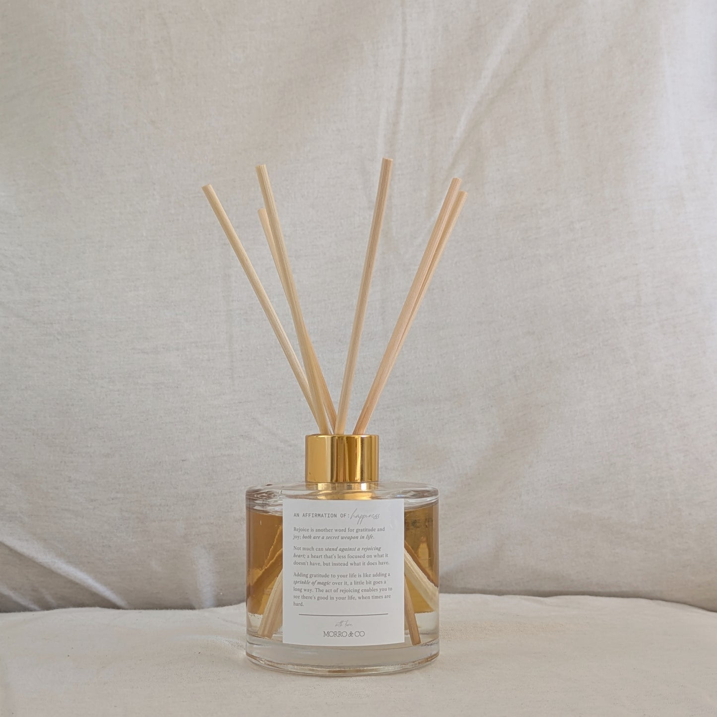Reed eco reed diffuser with gold cap, affirmation label and clear container on a neutral background
