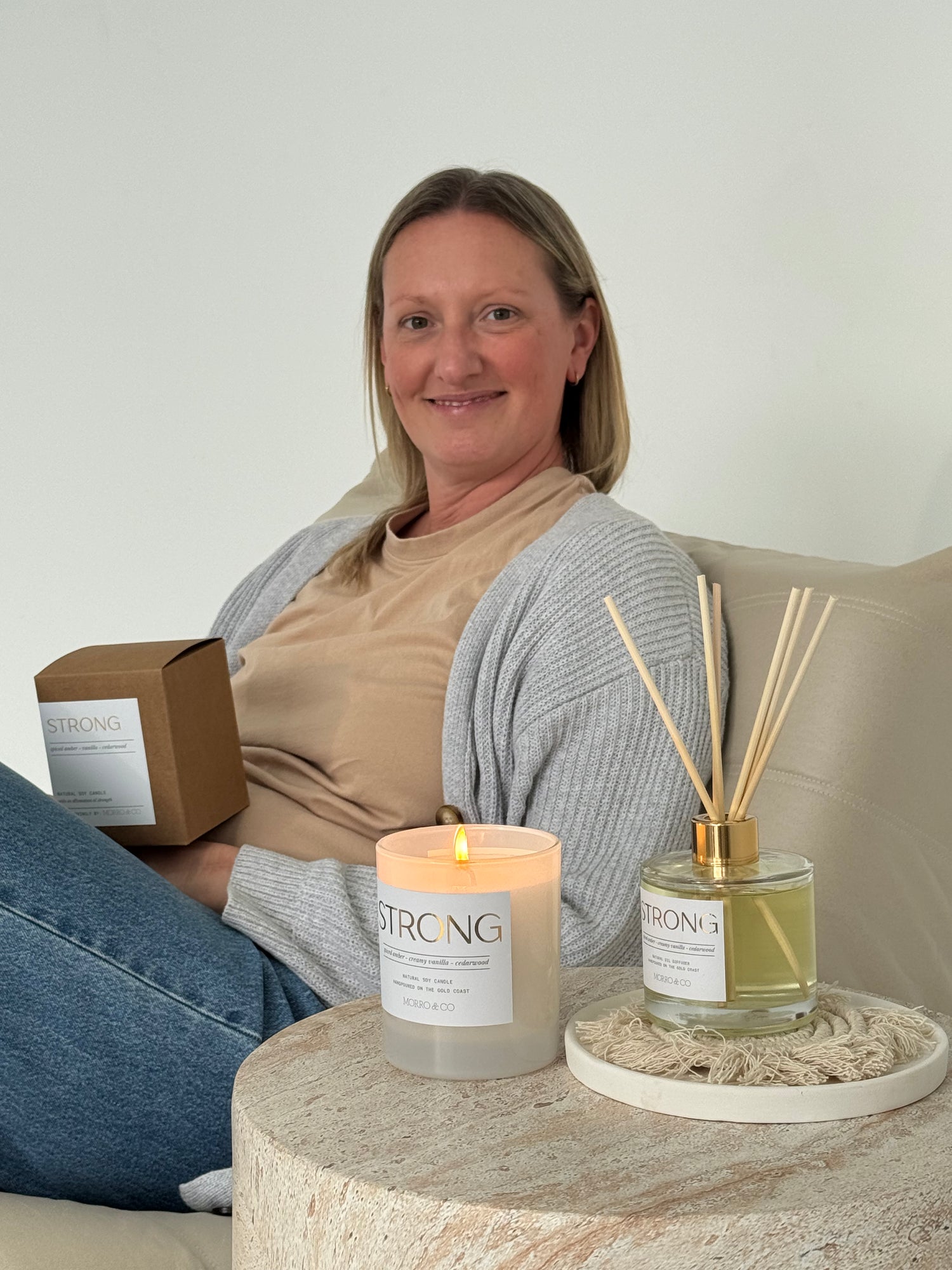 Woman sitting on a couch with a candle, diffuser, and box labeled 'STRONG' on a small table.
