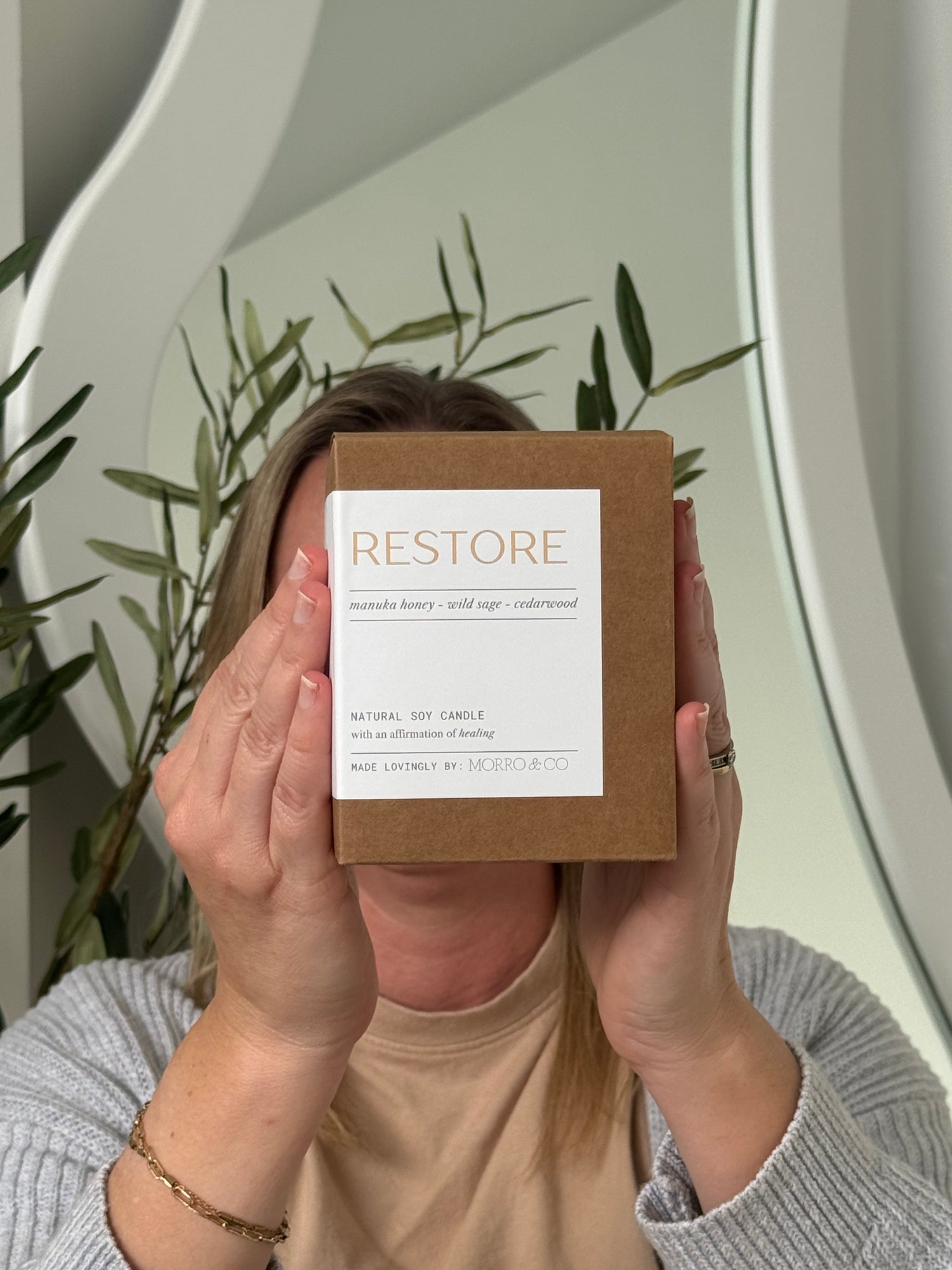 Person holding a 'Restore' candle box in front of their face with a neutral background