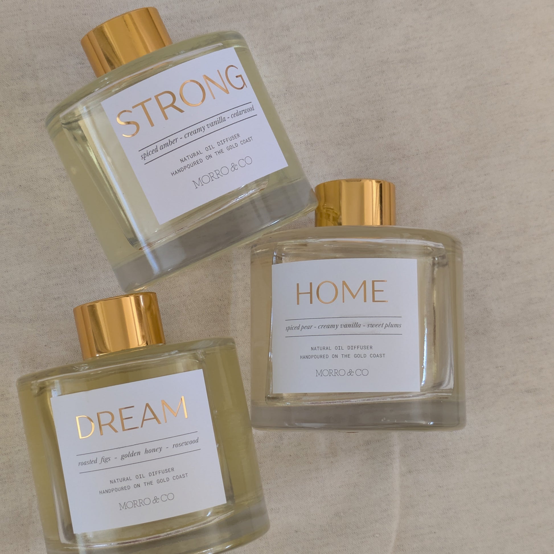 Three natural eco reed diffuser bottles labeled 'STRONG', 'HOME', and 'DREAM' on a beige surface.