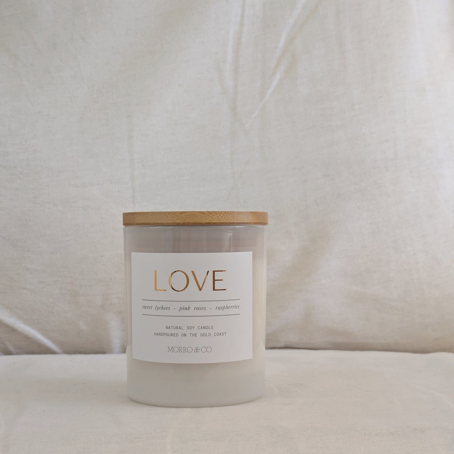 Front of candle with a wooden lid labeled with a gold label 'LOVE' on a beige fabric background