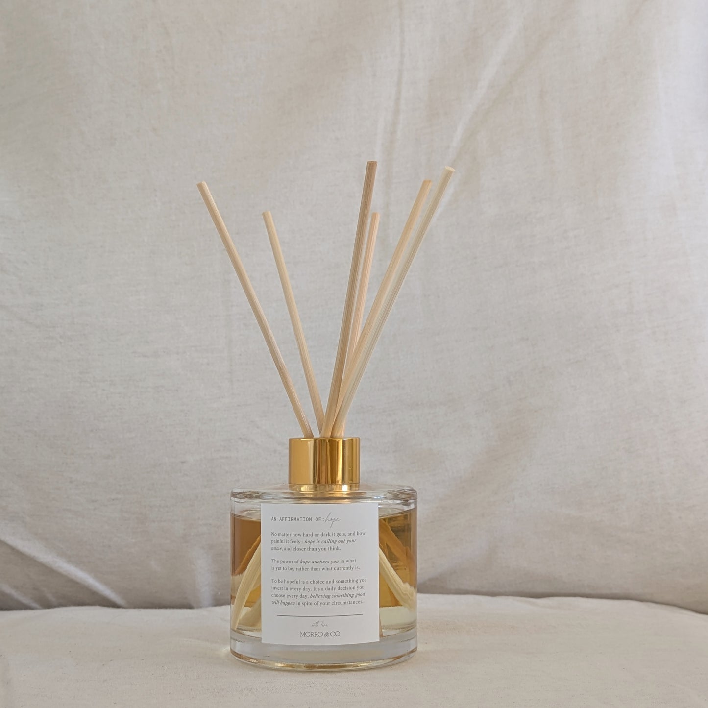 Natural eco reed diffuser with gold cap, affirmation label and clear bottle on a neutral background