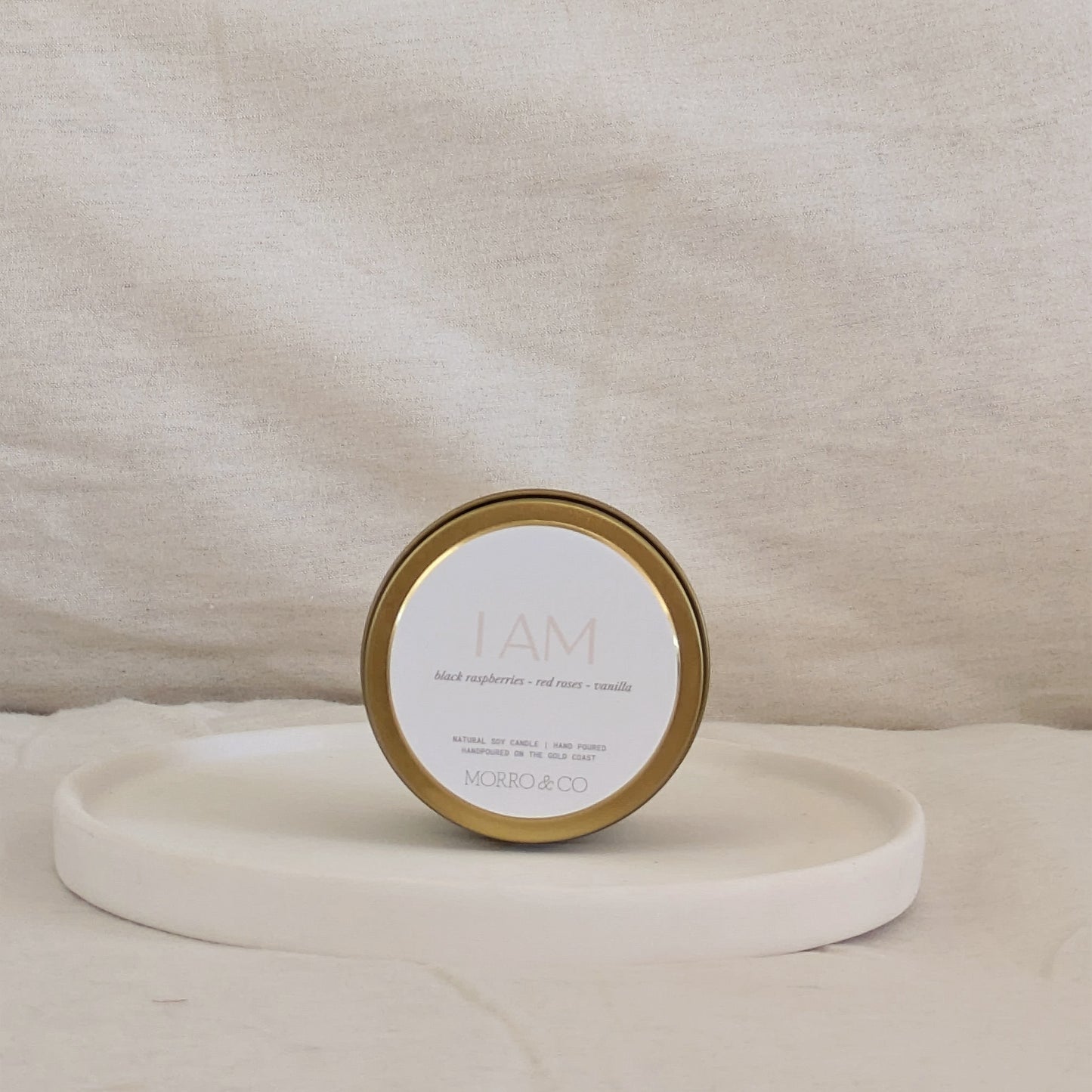 Gold candle travel tin with gold lid and labelled 'I AM' on a beige surface