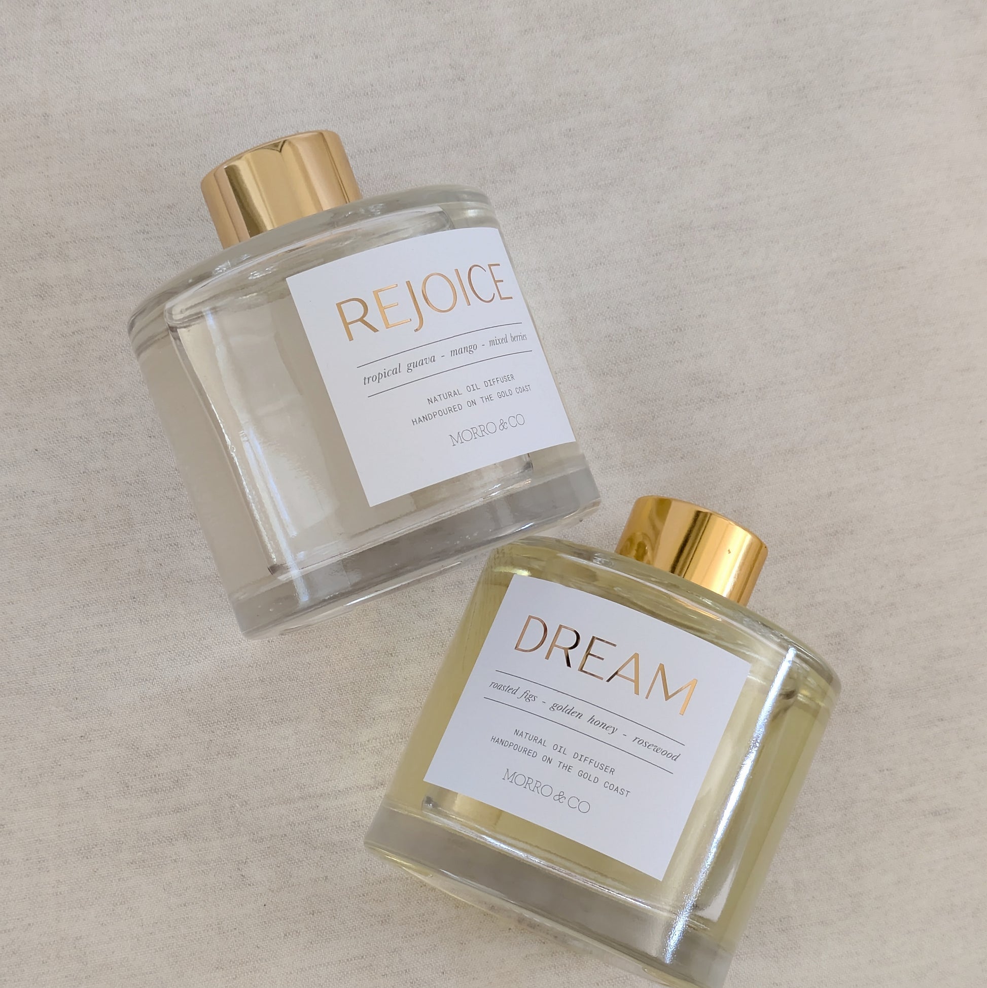Two eco reed diffusers bottles labeled 'Rejoice' and 'Dream' on a beige background