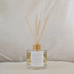 Eco reed diffuser with gold cap, affirmation label and clear bottle on a neutral background