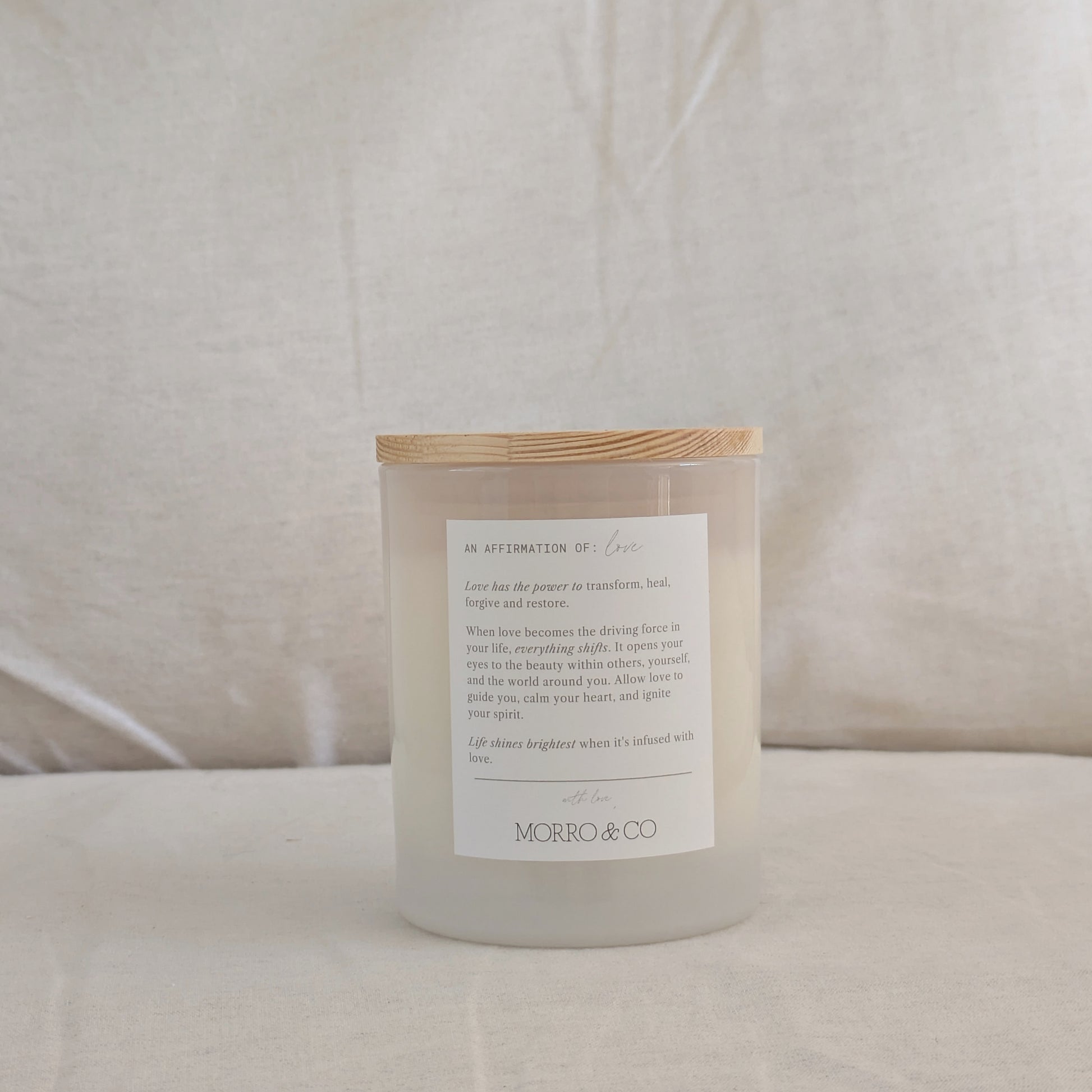 Back of LOVE candle with a wooden lid and an affirmation label on a neutral background