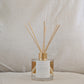 Reed diffuser with gold cap, affirmation label and clear container on a neutral background
