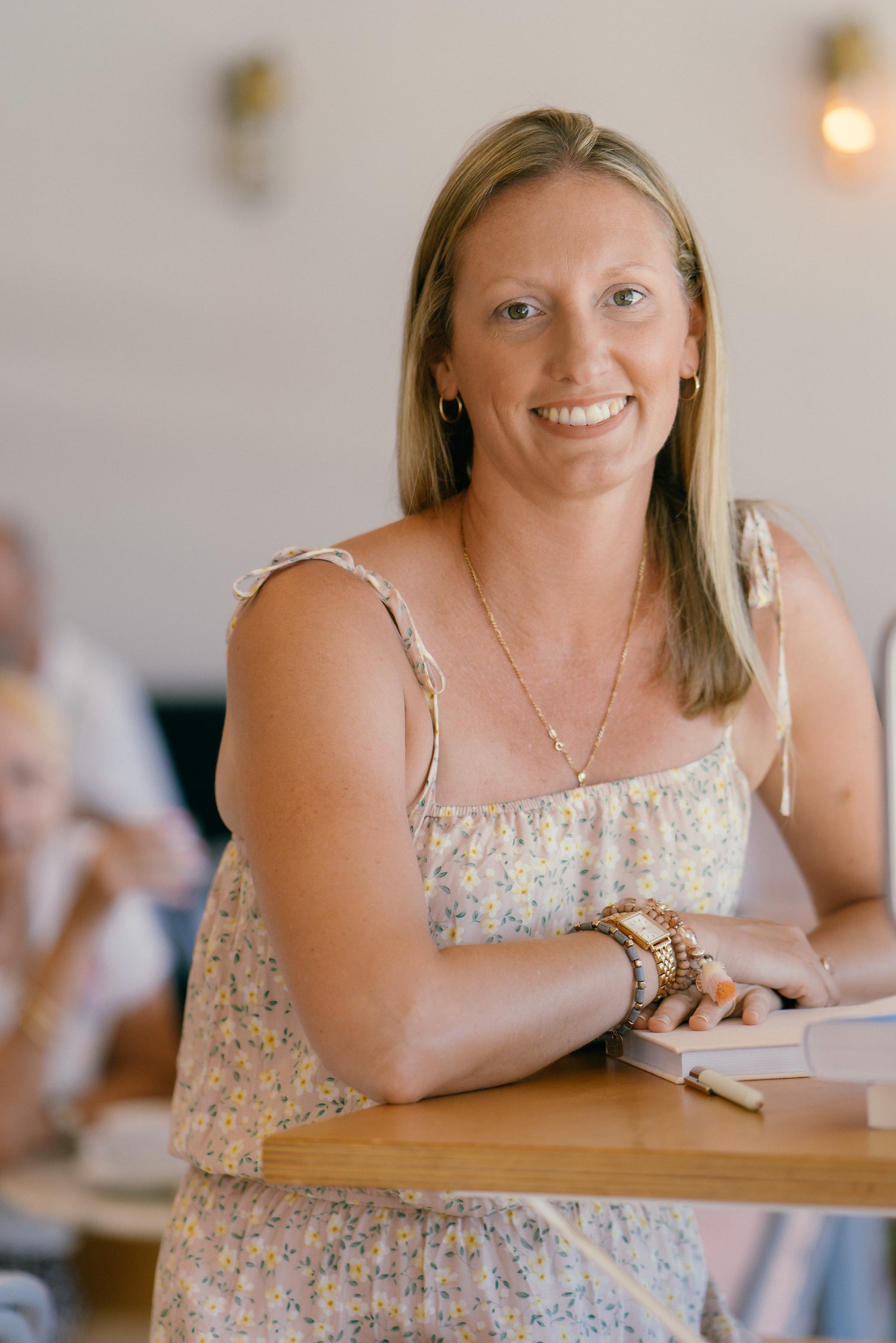 A smiling blonde woman wearing a light beige floral spaghetti-strap dress sits at a wooden table in a cafe or restaurant setting.