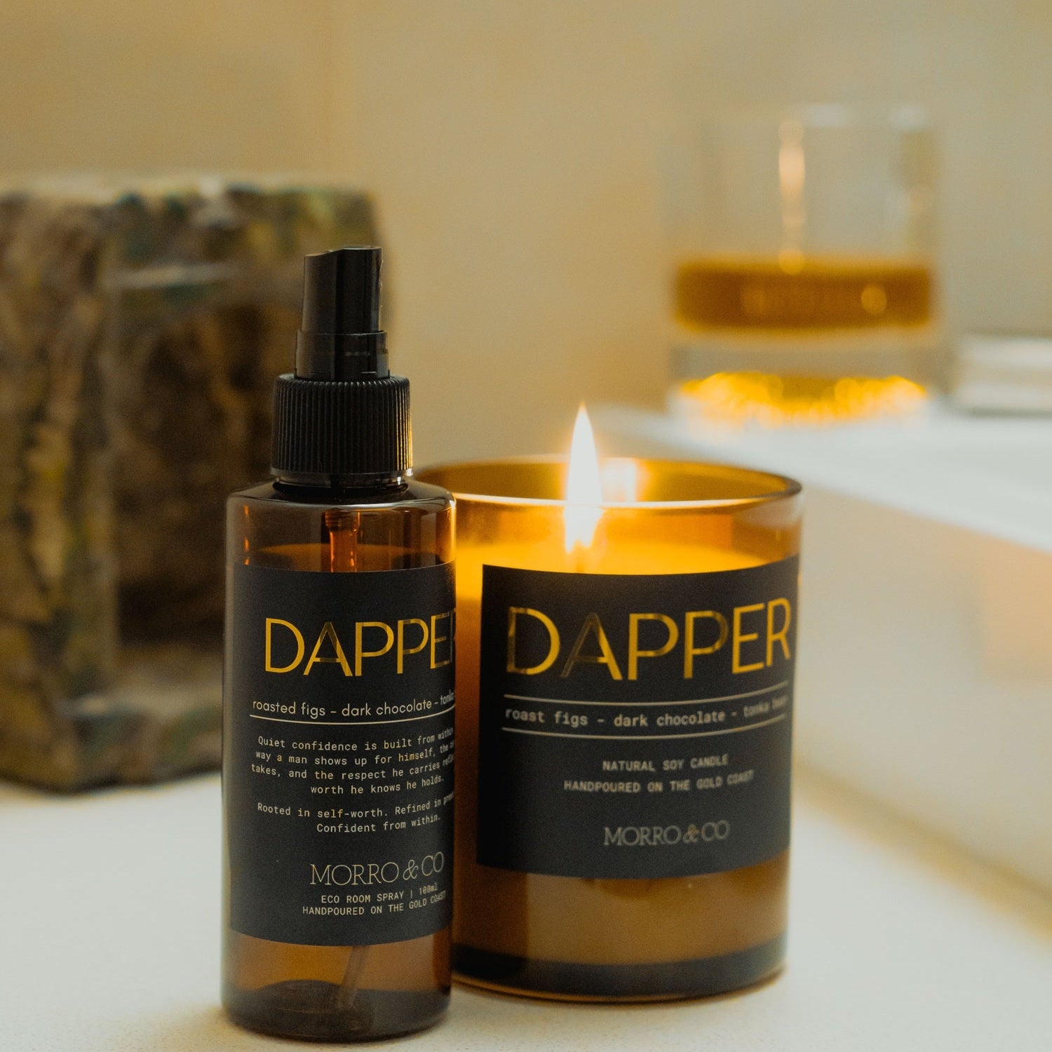 A Dapper Morbilloco natural soap and body care set, featuring a dark amber spray bottle and a matching lit candle in a glass jar, styled on a bathroom counter with a blurred glass of amber liquid in the background.