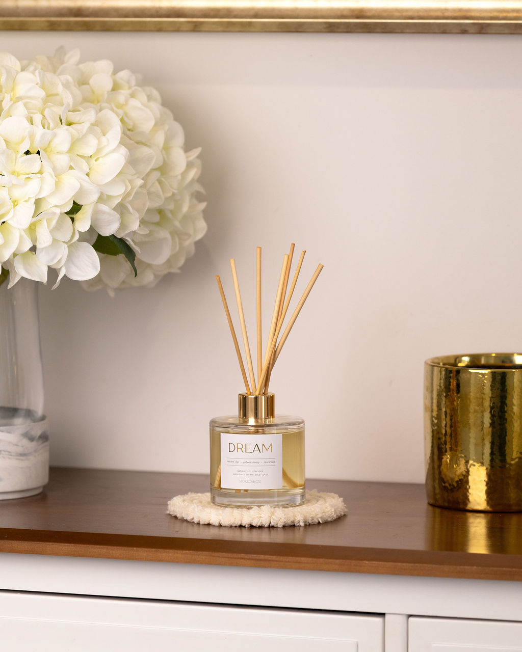 Diffuser with reeds on a table next to a vase of white flowers and a gold candle holder.
