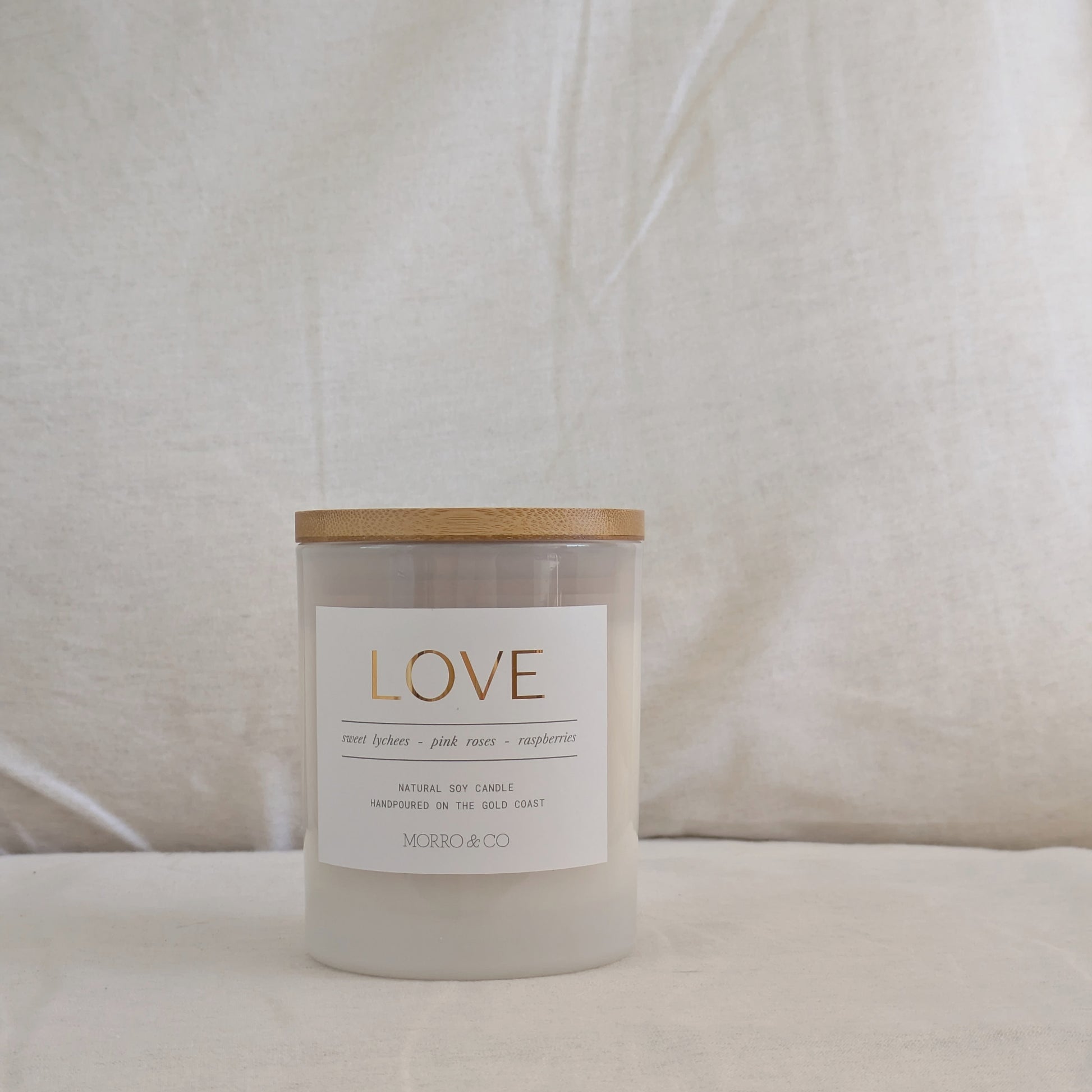 Front of candle with a wooden lid labeled with a gold label 'LOVE' on a beige fabric background