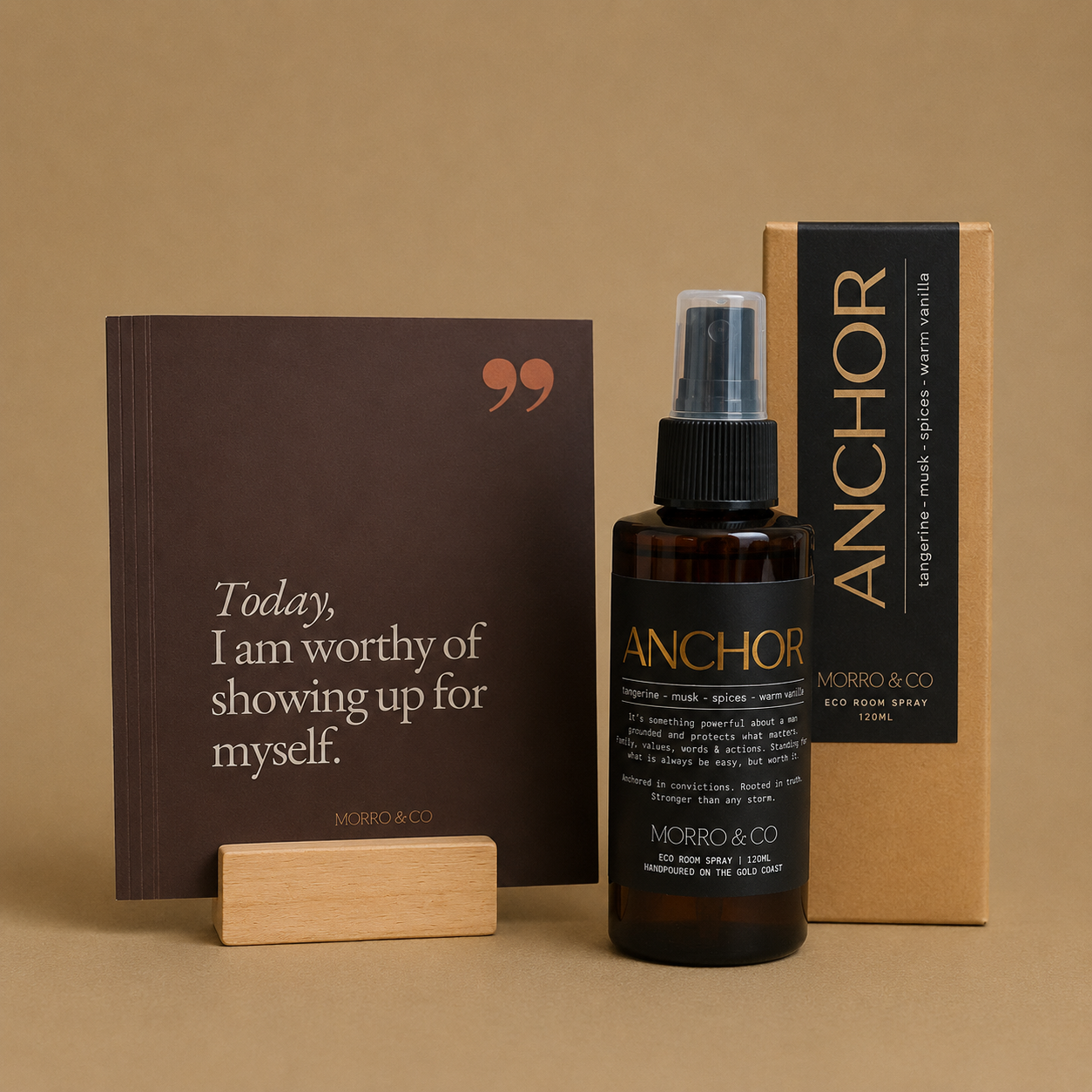 An amber glass spray bottle labeled ANCHOR MORRO & CO stands in front of its kraft paper packaging box, with a dark brown quote card and small wooden blocks on a warm beige background.