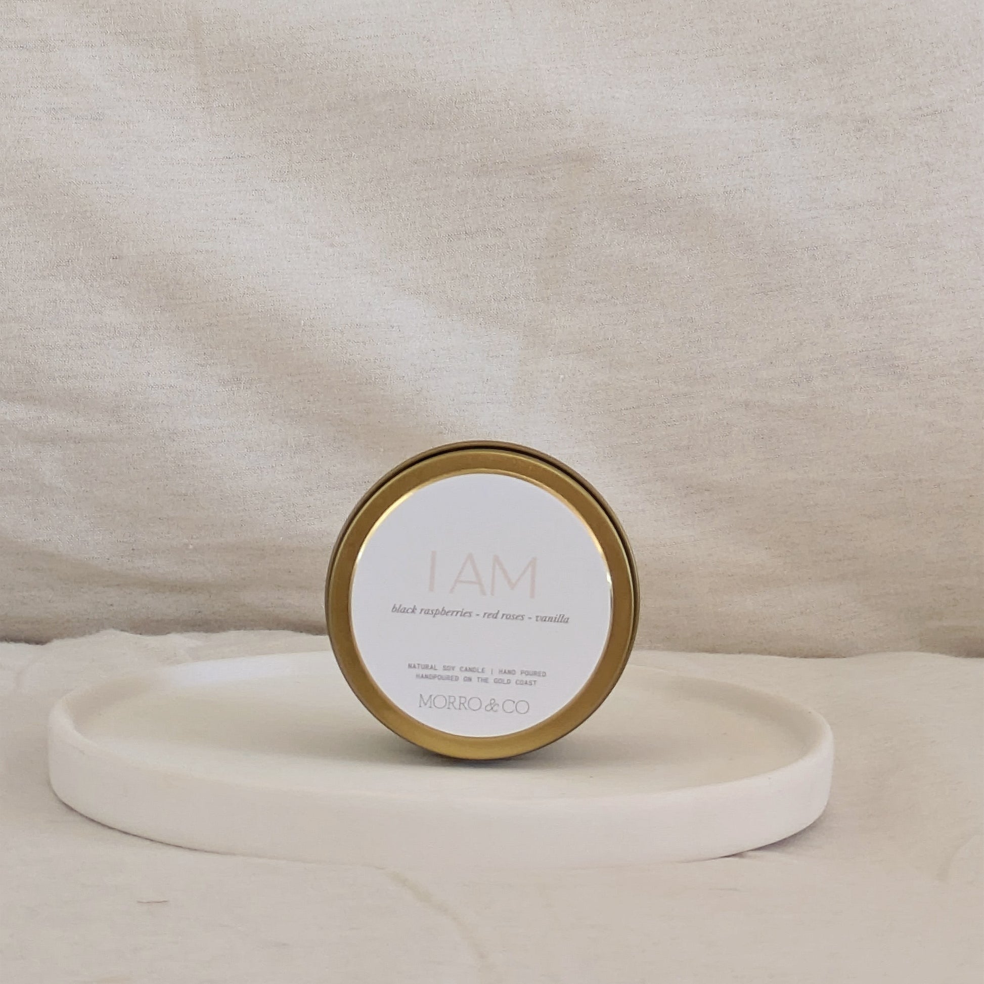 Gold candle travel tin with gold lid and labelled 'I AM' on a beige surface