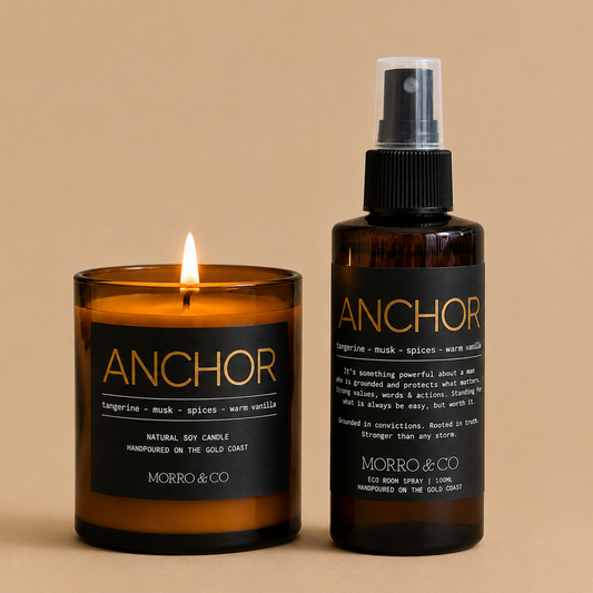 A lit amber glass candle jar and a matching dark amber spray bottle with black labels, both branded ANCHOR by MORRO & CO, set against a warm beige background.