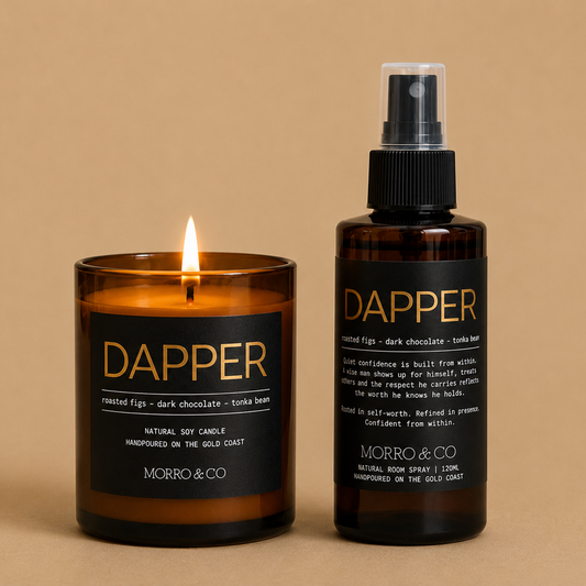 A candle and a matching room spray set from MORRO & CO, both labeled 'DAPPER' in gold lettering on black packaging, set against a solid beige background. The candle is lit, showing a warm amber glass body and a small flame, while the room spray is in an amber bottle with a black spray cap.