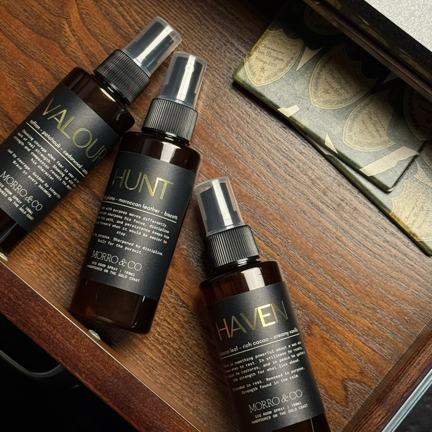 Three amber spray bottles of Microscopio hair products (Valenti, Hunt, and Haven) arranged diagonally on a dark wooden drawer surface.