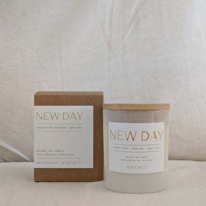 Candle labeled 'New Day' with packaging on a neutral background