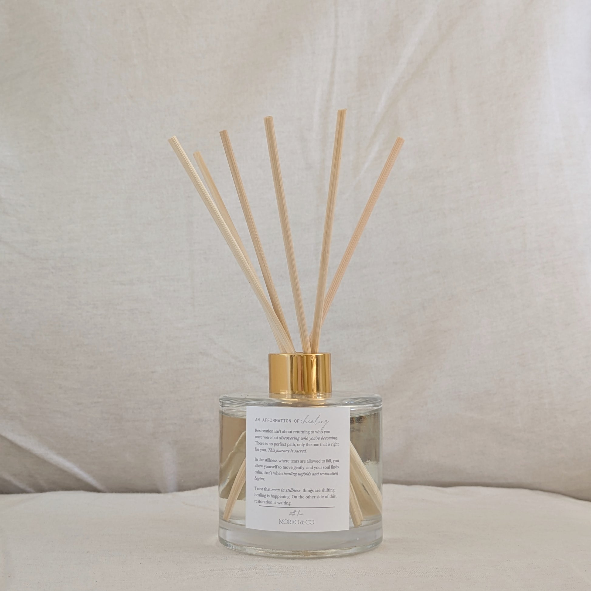 Reed diffuser with wooden sticks in a glass bottle with a gold cap and affirmation label on a neutral background