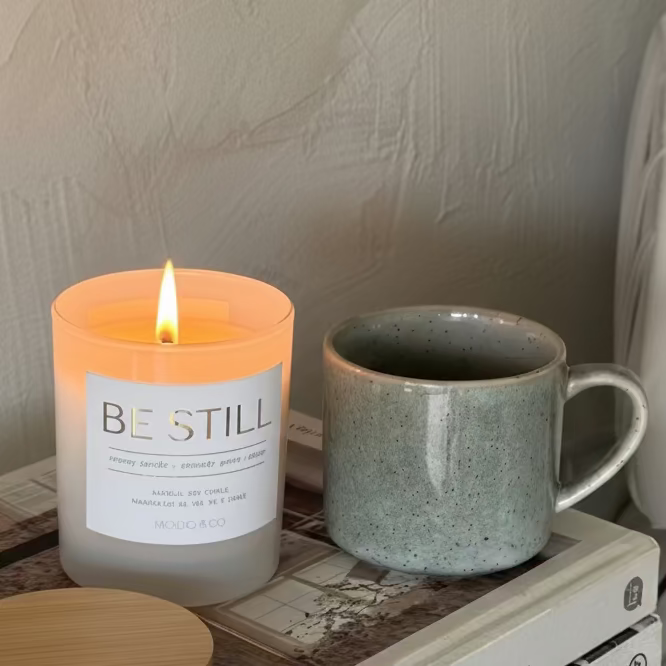 Candle labeled 'Be Still' on a wooden tray with a mug and coaster, against a neutral wall.