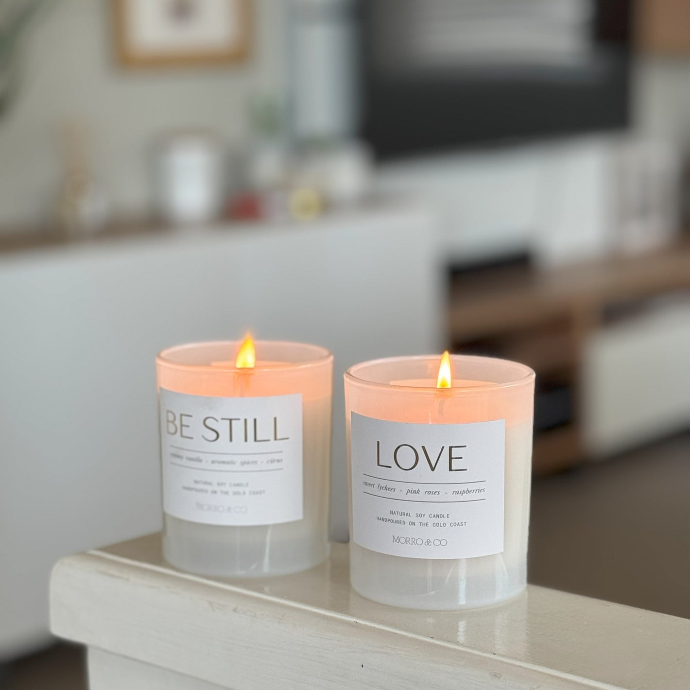 Two lit candles with 'BE STILL' and 'LOVE' labels on a surface in a blurred indoor setting.