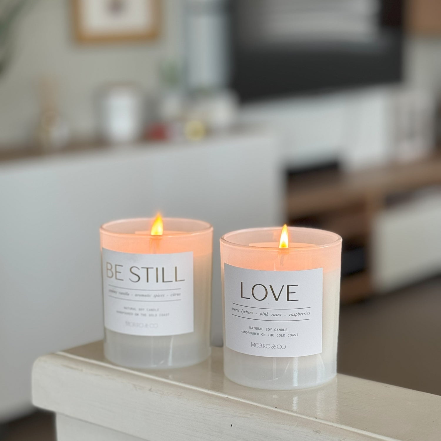 Two lit candles with 'BE STILL' and 'LOVE' labels on a surface in a blurred indoor setting.