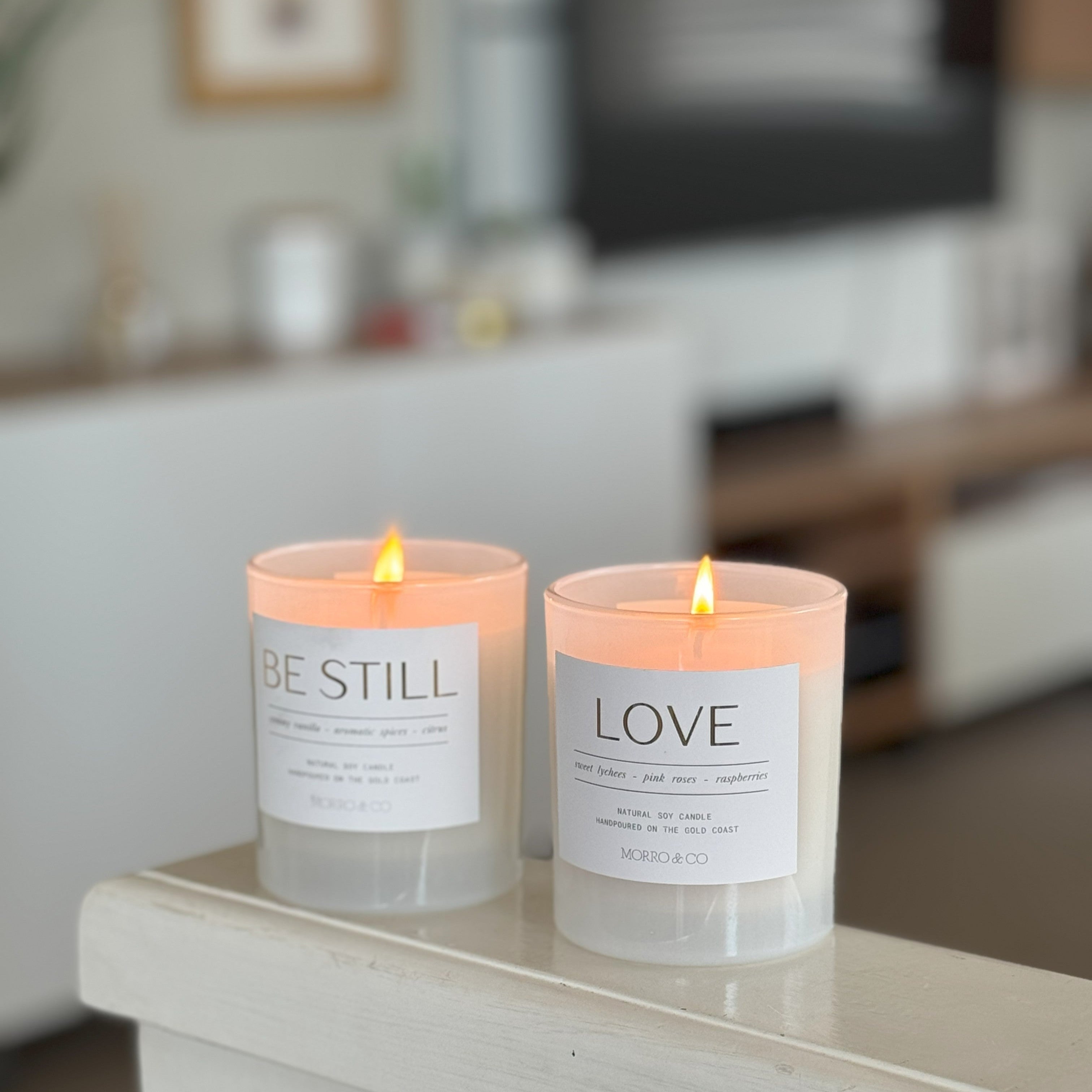 Two lit candles with 'BE STILL' and 'LOVE' labels on a surface in a blurred indoor setting.