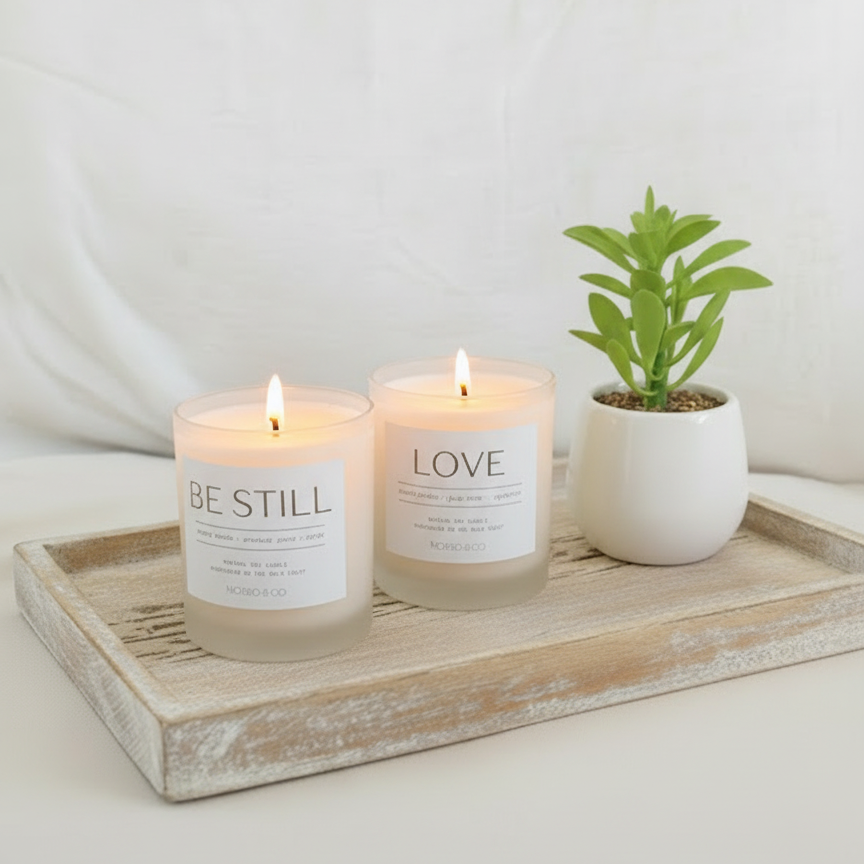 Two candles labeled 'Be Still' and 'Love' on a wooden tray with a small plant.