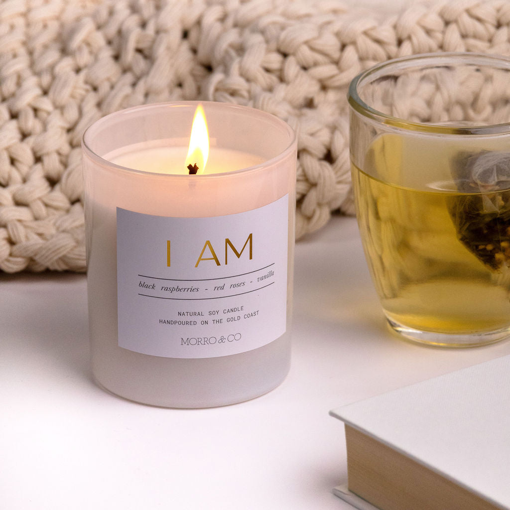 Candle with a label reading 'I AM' next to a glass of tea and a textured woven basket on a light surface.