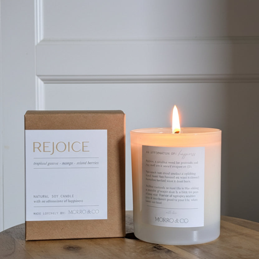 Candle and packaging labeled 'Rejoice' on a wooden surface with a neutral background